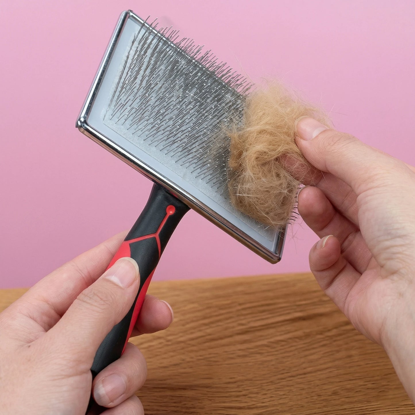 Mains retirant une touffe de poils dorés des picots métalliques d'une brosse carde professionnelle sur un fond rose.